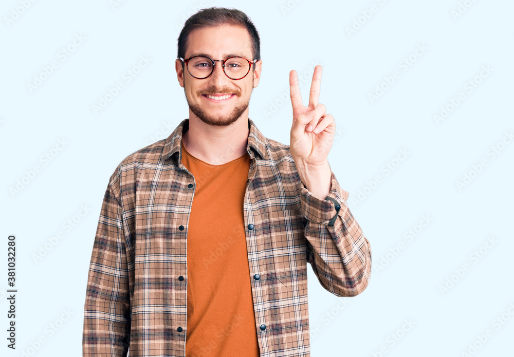 Young handsome caucasian man wearing casual clothes and glasses smiling with happy face winking at the camera doing victory sign. number two. Young handsome caucasian man wearing casual clothes and glasses smiling with happy face winking at the camera doing victory sign. number two.
