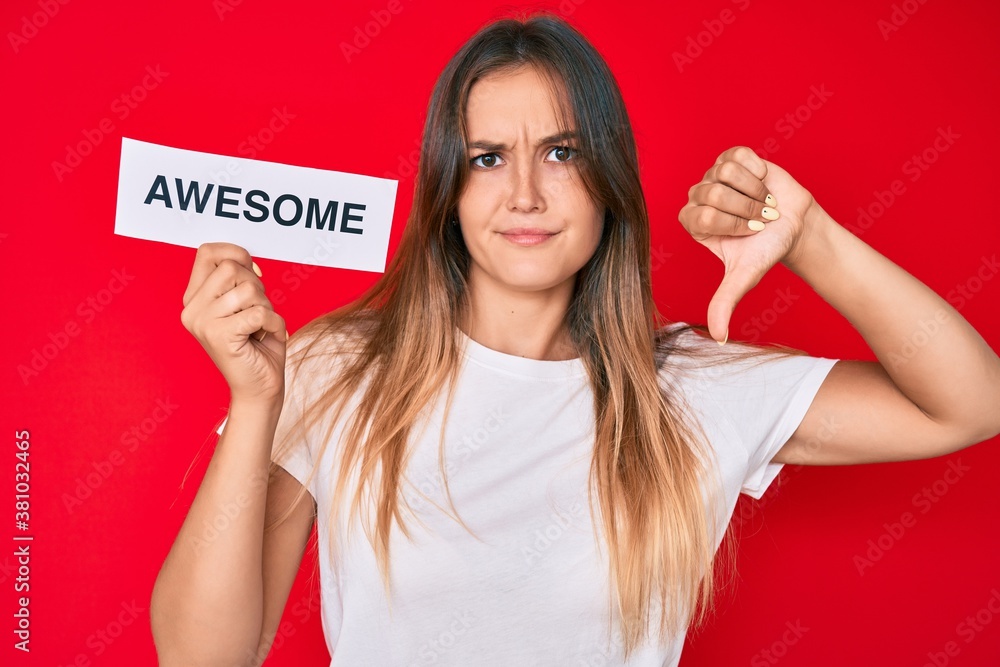 Beautiful caucasian woman holding awesome banner with angry face, negative sign showing dislike with thumbs down, rejection concept Beautiful caucasian woman holding awesome banner with angry face, negative sign showing dislike with thumbs down, rejection concept