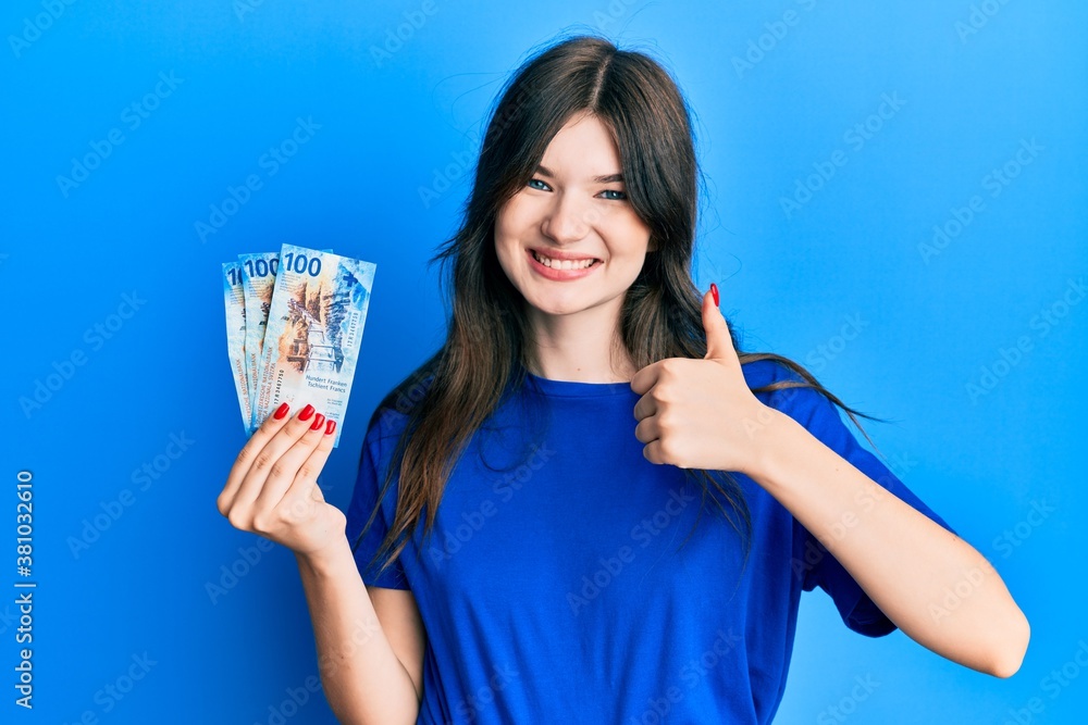 Young beautiful caucasian girl holding 100 swiss franc banknotes smiling happy and positive, thumb up doing excellent and approval sign Young beautiful caucasian girl holding 100 swiss franc banknotes smiling happy and positive, thumb up doing excellent and approval sign