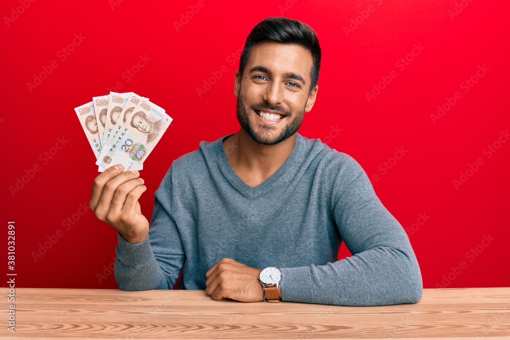 Handsome hispanic man holding yuan chinese banknotes looking positive and happy standing and smiling with a confident smile showing teeth Handsome hispanic man holding yuan chinese banknotes looking positive and happy standing and smiling with a confident smile showing teeth