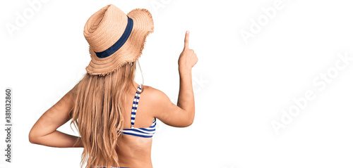 Young beautiful blonde woman wearing bikini and hat posing backwards pointing ahead with finger hand photo