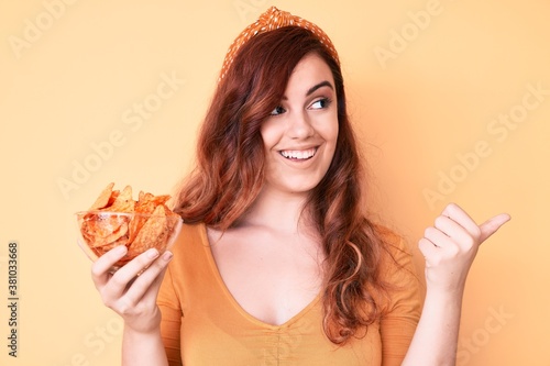Young beautiful woman holding nachos potato chips pointing thumb up to the side smiling happy with open mouth