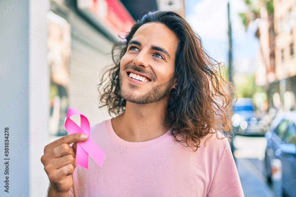 Young hispanic man smiling happy holding breast cancer pink ribbon at city.