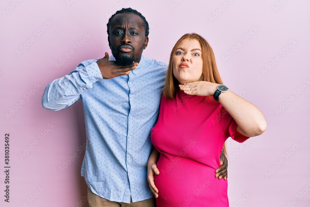 Obraz premium Young interracial couple expecting a baby, touching pregnant belly cutting throat with hand as knife, threaten aggression with furious violence