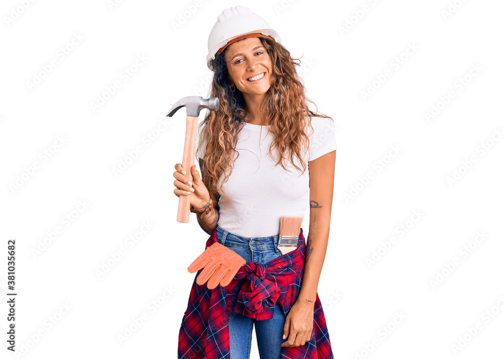 Young hispanic woman with tattoo wearing hardhat and builder clothes holding hammer looking positive and happy standing and smiling with a confident smile showing teeth Young hispanic woman with tattoo wearing hardhat and builder clothes holding hammer looking positive and happy standing and smiling with a confident smile showing teeth
