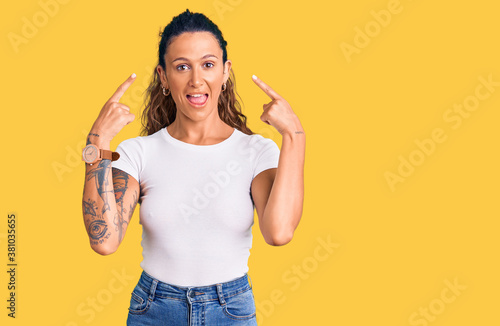 Young hispanic woman with tattoo wearing casual white tshirt smiling pointing to head with both hands finger, great idea or thought, good memory