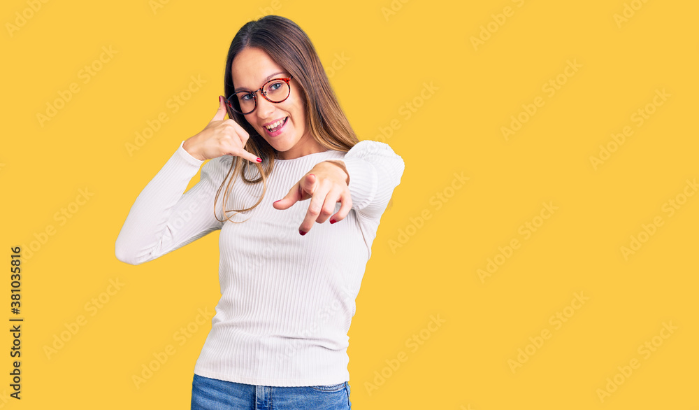 Beautiful brunette young woman wearing casual white sweater and glasses smiling doing talking on the telephone gesture and pointing to you. call me. Beautiful brunette young woman wearing casual white sweater and glasses smiling doing talking on the telephone gesture and pointing to you. call me.