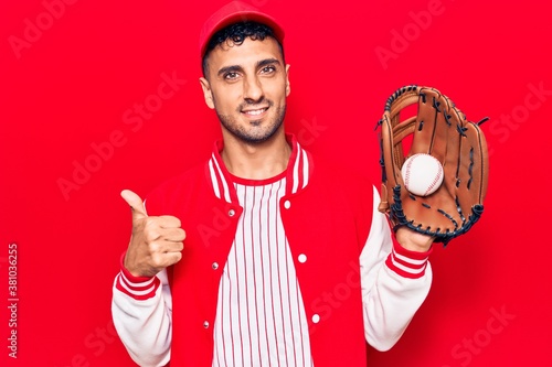 Young hispanic man wearing baseball uniform holding golve and ball smiling happy and positive, thumb up doing excellent and approval sign photo