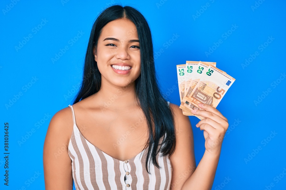 Young beautiful asian girl holding euro banknotes looking positive and happy standing and smiling with a confident smile showing teeth Young beautiful asian girl holding euro banknotes looking positive and happy standing and smiling with a confident smile showing teeth