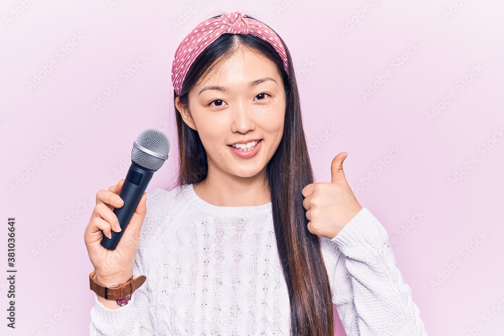Young beautiful chinese woman singing song using microphone smiling happy and positive, thumb up doing excellent and approval sign Young beautiful chinese woman singing song using microphone smiling happy and positive, thumb up doing excellent and approval sign