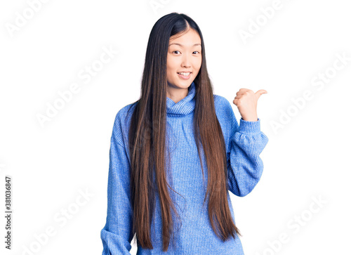 Young beautiful chinese woman wearing casual turtleneck sweater smiling with happy face looking and pointing to the side with thumb up. © Krakenimages.com