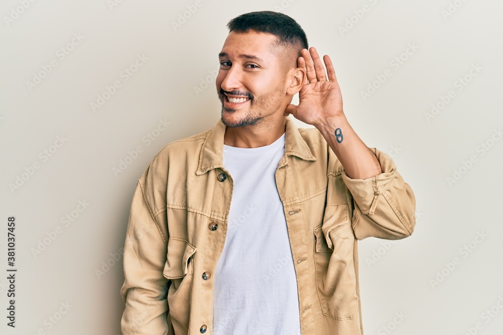Handsome man with tattoos wearing casual brown denim jacket smiling with hand over ear listening an hearing to rumor or gossip. deafness concept. Handsome man with tattoos wearing casual brown denim jacket smiling with hand over ear listening an hearing to rumor or gossip. deafness concept.