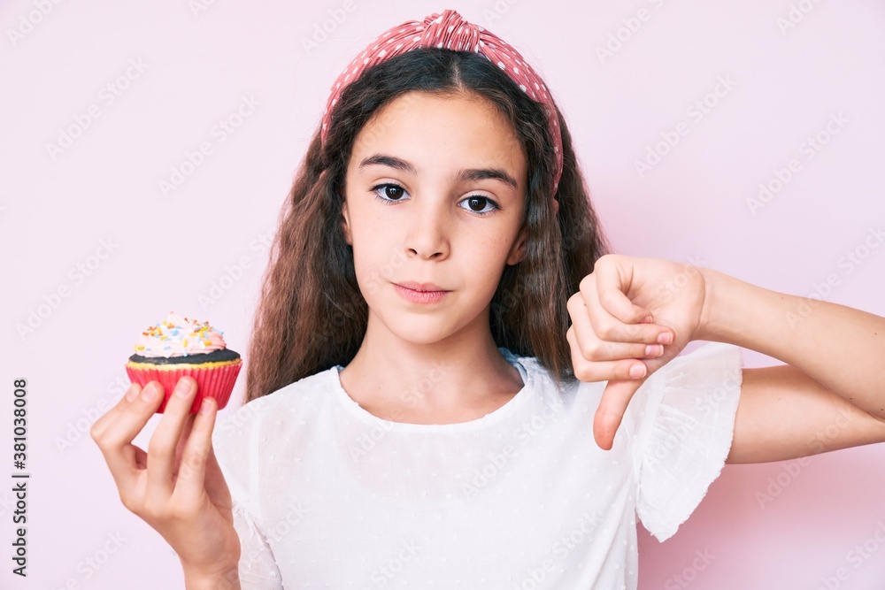 Cute hispanic child girl holding cupcake with angry face, negative sign showing dislike with thumbs down, rejection concept Cute hispanic child girl holding cupcake with angry face, negative sign showing dislike with thumbs down, rejection concept