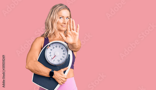 Middle age fit blonde woman wearing sports clothes holding weighing machine with open hand doing stop sign with serious and confident expression, defense gesture