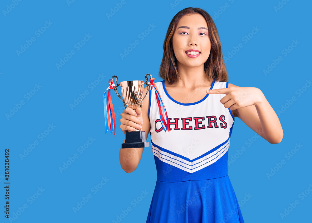 Youn beautiful asian girl wearing cheerleader uniform holding champion trophy smiling happy pointing with hand and finger Youn beautiful asian girl wearing cheerleader uniform holding champion trophy smiling happy pointing with hand and finger