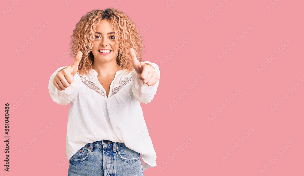 Young blonde woman with curly hair wearing elegant summer shirt approving doing positive gesture with hand, thumbs up smiling and happy for success. winner gesture. Young blonde woman with curly hair wearing elegant summer shirt approving doing positive gesture with hand, thumbs up smiling and happy for success. winner gesture.