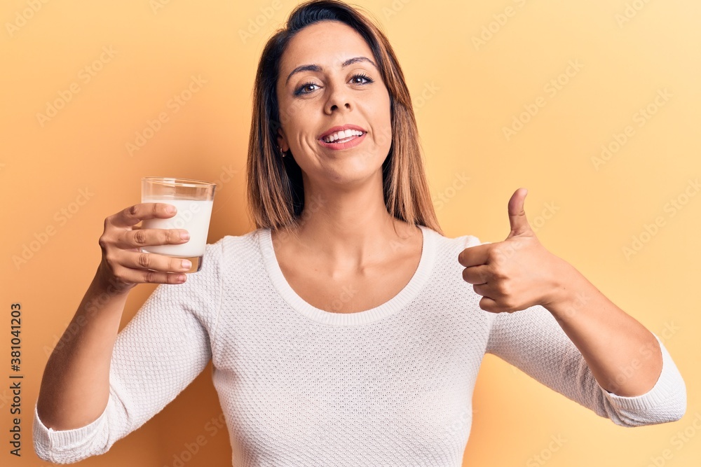 Young beautiful woman holding glass of milk smiling happy and positive, thumb up doing excellent and approval sign Young beautiful woman holding glass of milk smiling happy and positive, thumb up doing excellent and approval sign