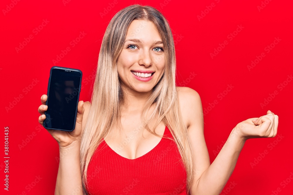 Young beautiful blonde girl holding smartphone showing screen screaming proud, celebrating victory and success very excited with raised arm Young beautiful blonde girl holding smartphone showing screen screaming proud, celebrating victory and success very excited with raised arm