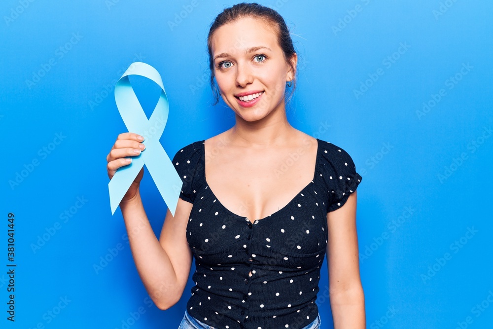 Young beautiful blonde woman holding blue ribbon looking positive and happy standing and smiling with a confident smile showing teeth Young beautiful blonde woman holding blue ribbon looking positive and happy standing and smiling with a confident smile showing teeth