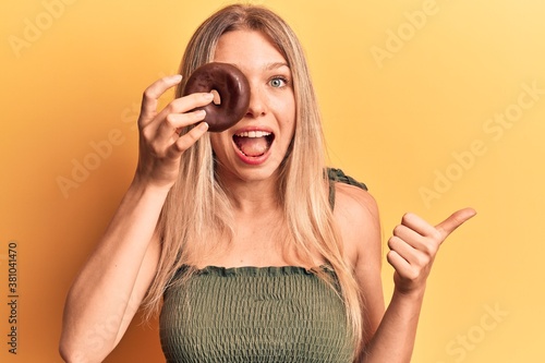 Young blonde woman holding donut pointing thumb up to the side smiling happy with open mouth