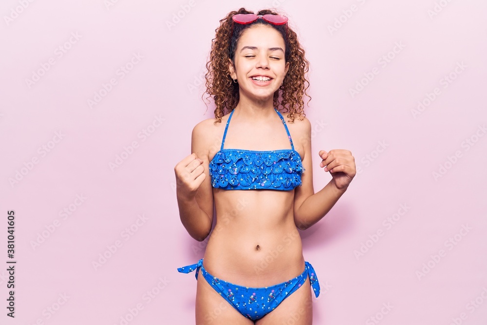 Naklejka premium Beautiful kid girl with curly hair wearing bikini and sunglasses very happy and excited doing winner gesture with arms raised, smiling and screaming for success. celebration concept.