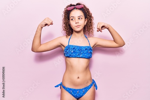 Beautiful kid girl with curly hair wearing bikini and sunglasses showing arms muscles smiling proud. fitness concept.