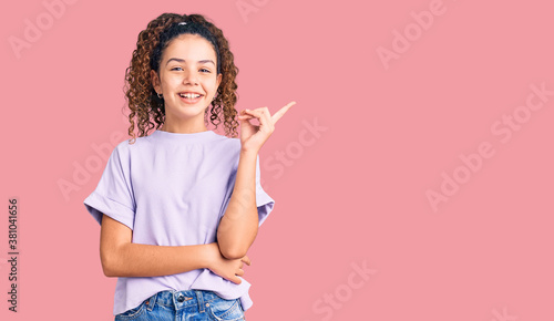 Beautiful kid girl with curly hair wearing casual clothes with a big smile on face, pointing with hand and finger to the side looking at the camera.