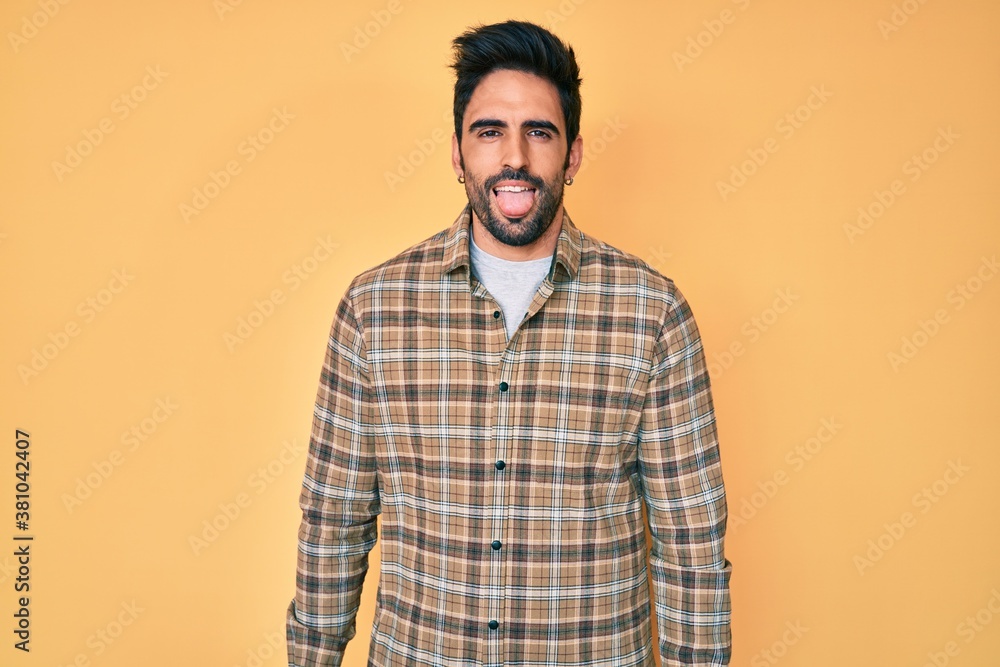 Handsome hispanic man with beard wearing casual clothes sticking tongue out happy with funny expression. emotion concept. Handsome hispanic man with beard wearing casual clothes sticking tongue out happy with funny expression. emotion concept.