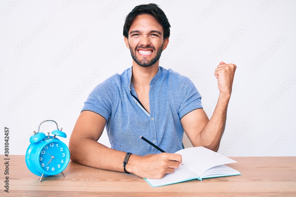 Handsome hispanic man sitting on the table stuying for university screaming proud, celebrating victory and success very excited with raised arms Handsome hispanic man sitting on the table stuying for university screaming proud, celebrating victory and success very excited with raised arms
