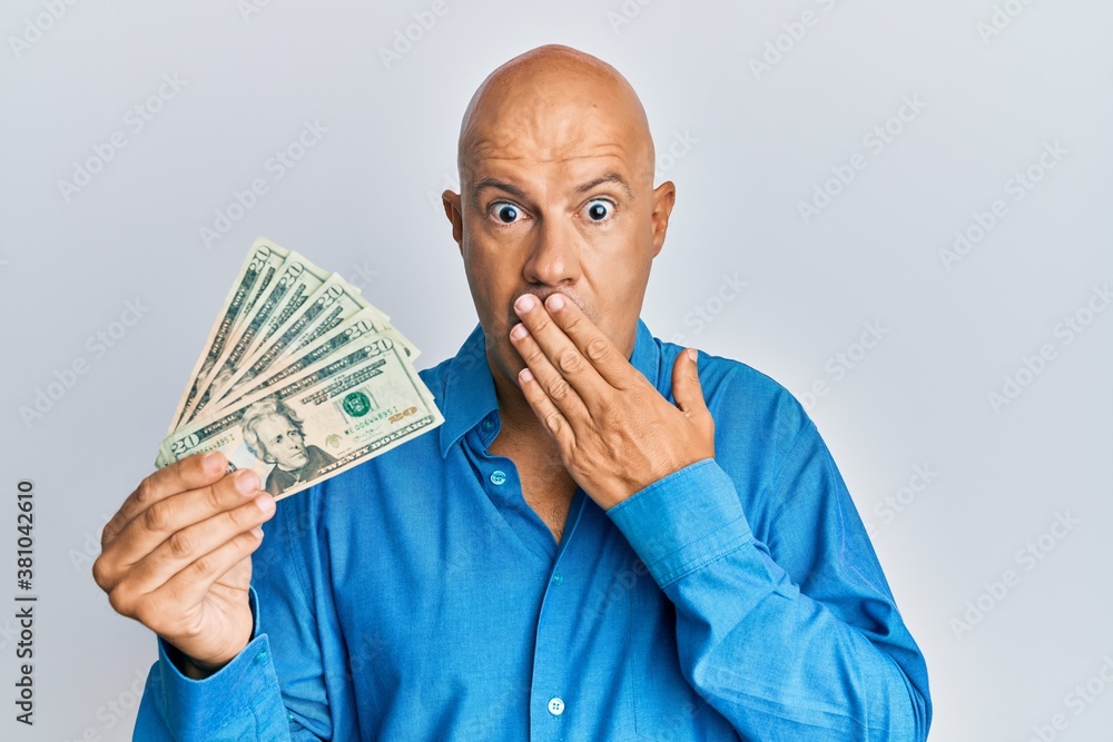 Middle age bald man holding 20 dollars banknote covering mouth with hand, shocked and afraid for mistake. surprised expression Middle age bald man holding 20 dollars banknote covering mouth with hand, shocked and afraid for mistake. surprised expression