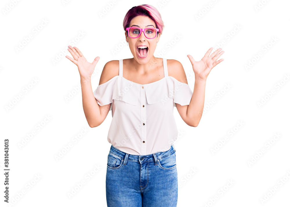 Young beautiful woman with pink hair wearing casual clothes and glasses celebrating crazy and amazed for success with arms raised and open eyes screaming excited. winner concept Young beautiful woman with pink hair wearing casual clothes and glasses celebrating crazy and amazed for success with arms raised and open eyes screaming excited. winner concept