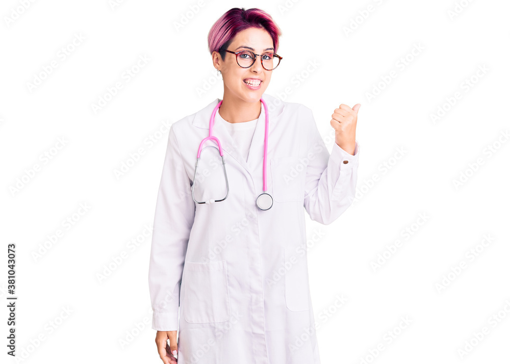 Young beautiful woman with pink hair wearing doctor uniform smiling with happy face looking and pointing to the side with thumb up. Young beautiful woman with pink hair wearing doctor uniform smiling with happy face looking and pointing to the side with thumb up.