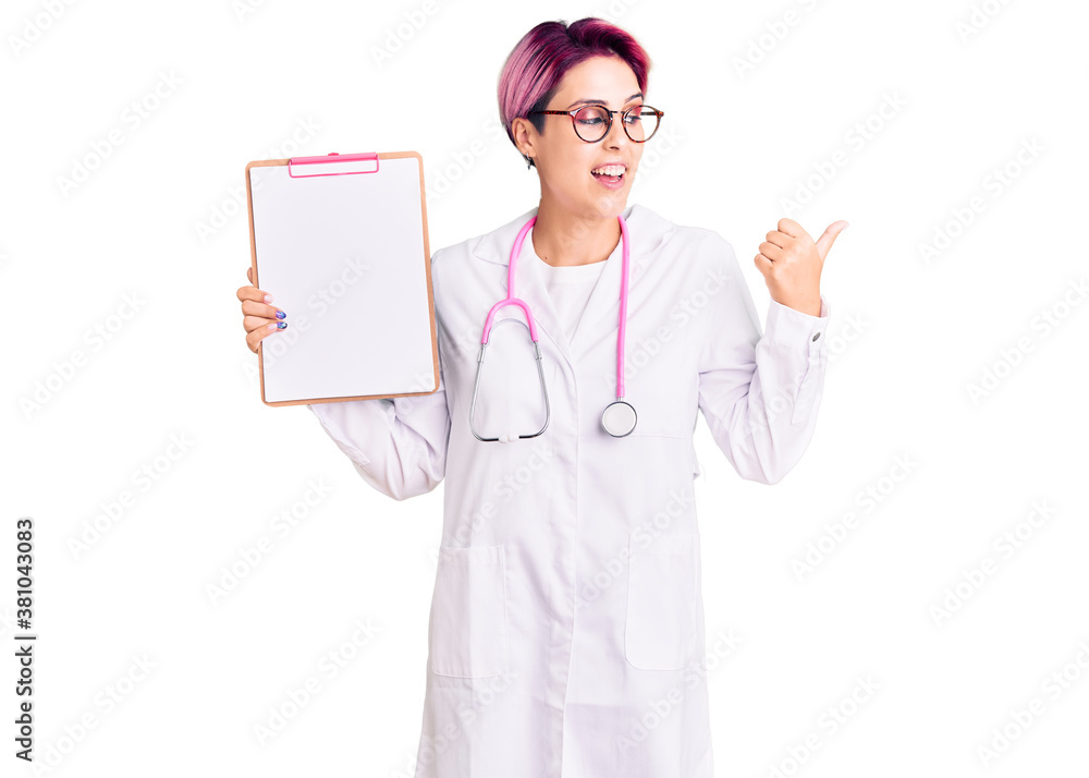 Young beautiful woman with pink hair wearing doctor stethoscope holding clipboard with medical report pointing thumb up to the side smiling happy with open mouth Young beautiful woman with pink hair wearing doctor stethoscope holding clipboard with medical report pointing thumb up to the side smiling happy with open mouth