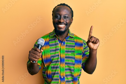 Handsome young black man singing song using microphone smiling with an idea or question pointing finger with happy face, number one