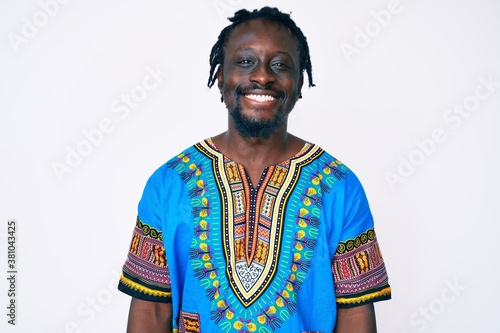 Young african american man with braids wearing traditional africa tshirt looking positive and happy standing and smiling with a confident smile showing teeth