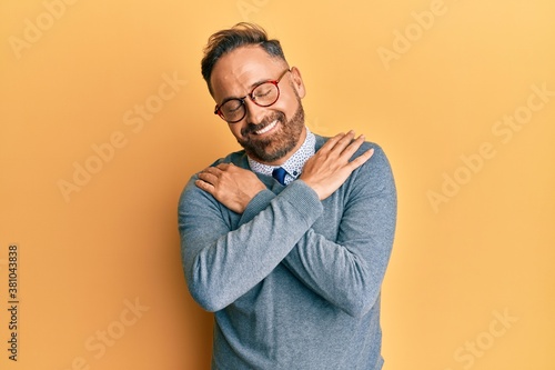 Handsome middle age man wearing glasses hugging oneself happy and positive, smiling confident. self love and self care © Krakenimages.com