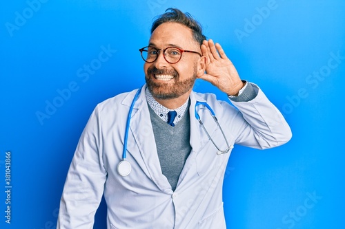Handsome middle age man wearing doctor uniform and stethoscope smiling with hand over ear listening an hearing to rumor or gossip. deafness concept.