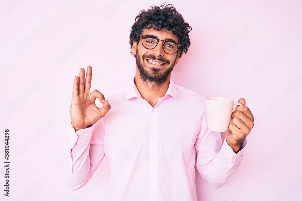 Handsome young man with curly hair and bear wearing glasses and drinking a cup of coffee doing ok sign with fingers, smiling friendly gesturing excellent symbol Handsome young man with curly hair and bear wearing glasses and drinking a cup of coffee doing ok sign with fingers, smiling friendly gesturing excellent symbol
