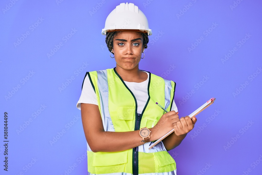Fototapeta premium Young african american woman with braids wearing safety helmet holding clipboard skeptic and nervous, frowning upset because of problem. negative person.