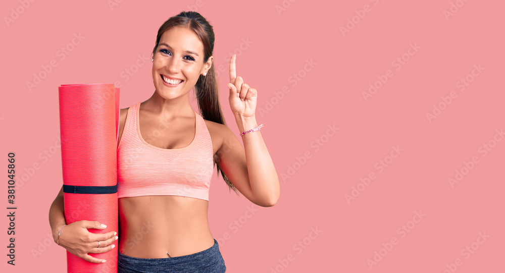 Young beautiful hispanic woman holding yoga mat surprised with an idea or question pointing finger with happy face, number one