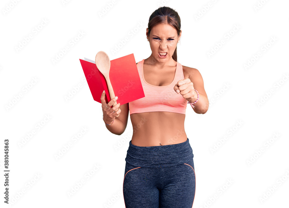 Young beautiful hispanic woman wearing gym clothes reading cooking recipe book holding spoon annoyed and frustrated shouting with anger, yelling crazy with anger and hand raised Young beautiful hispanic woman wearing gym clothes reading cooking recipe book holding spoon annoyed and frustrated shouting with anger, yelling crazy with anger and hand raised