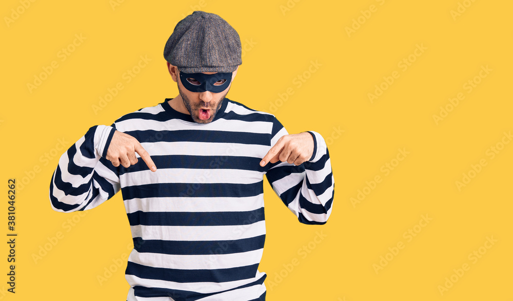 Young handsome man wearing burglar mask pointing down with fingers showing advertisement, surprised face and open mouth Young handsome man wearing burglar mask pointing down with fingers showing advertisement, surprised face and open mouth