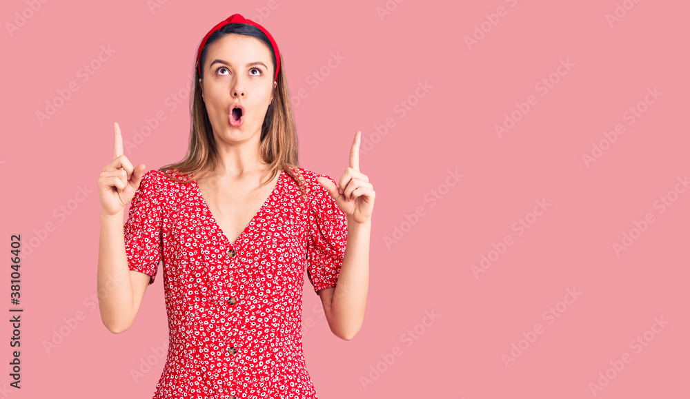 Young beautiful girl wearing dress and diadem amazed and surprised looking up and pointing with fingers and raised arms. Young beautiful girl wearing dress and diadem amazed and surprised looking up and pointing with fingers and raised arms.