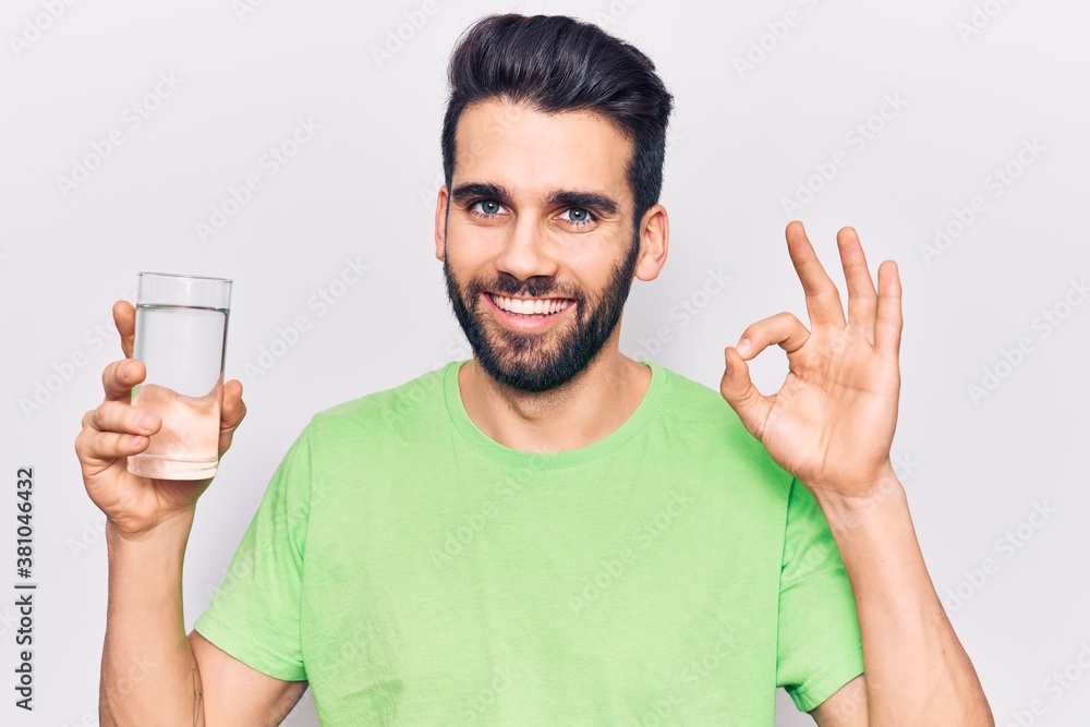 Young handsome man with beard drinking glass of water doing ok sign with fingers, smiling friendly gesturing excellent symbol Young handsome man with beard drinking glass of water doing ok sign with fingers, smiling friendly gesturing excellent symbol