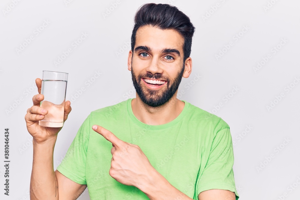 Young handsome man with beard drinking glass of water smiling happy pointing with hand and finger Young handsome man with beard drinking glass of water smiling happy pointing with hand and finger