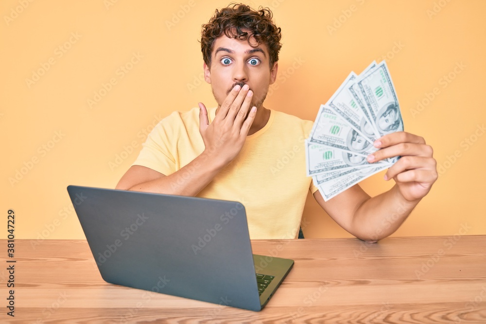 Young caucasian man with curly hair working using computer laptop holding usa dollars banknotes covering mouth with hand, shocked and afraid for mistake. surprised expression Young caucasian man with curly hair working using computer laptop holding usa dollars banknotes covering mouth with hand, shocked and afraid for mistake. surprised expression