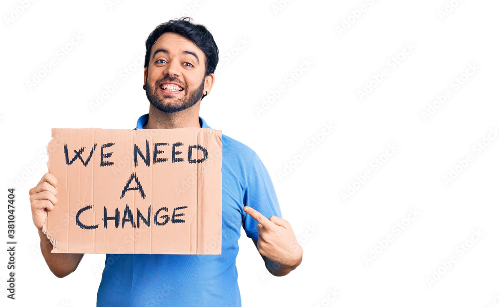 Young hispanic man holding we need a change banner smiling happy pointing with hand and finger Young hispanic man holding we need a change banner smiling happy pointing with hand and finger