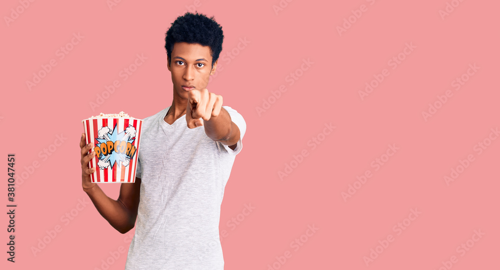Young african american man holding popcorn pointing with finger to the camera and to you, confident gesture looking serious Young african american man holding popcorn pointing with finger to the camera and to you, confident gesture looking serious
