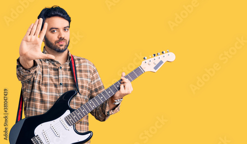 Young hispanic man playing electric guitar with open hand doing stop sign with serious and confident expression, defense gesture