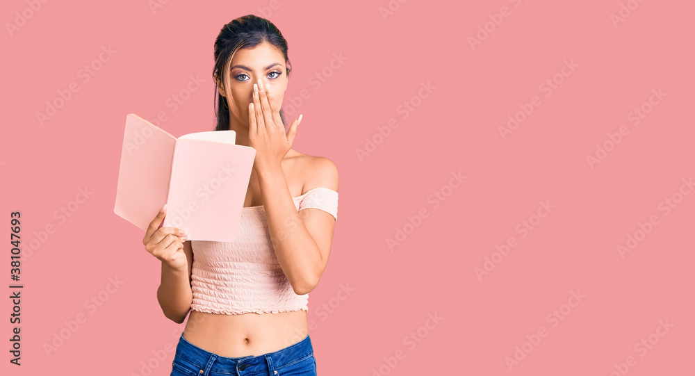 Naklejka premium Young beautiful woman holding book covering mouth with hand, shocked and afraid for mistake. surprised expression
